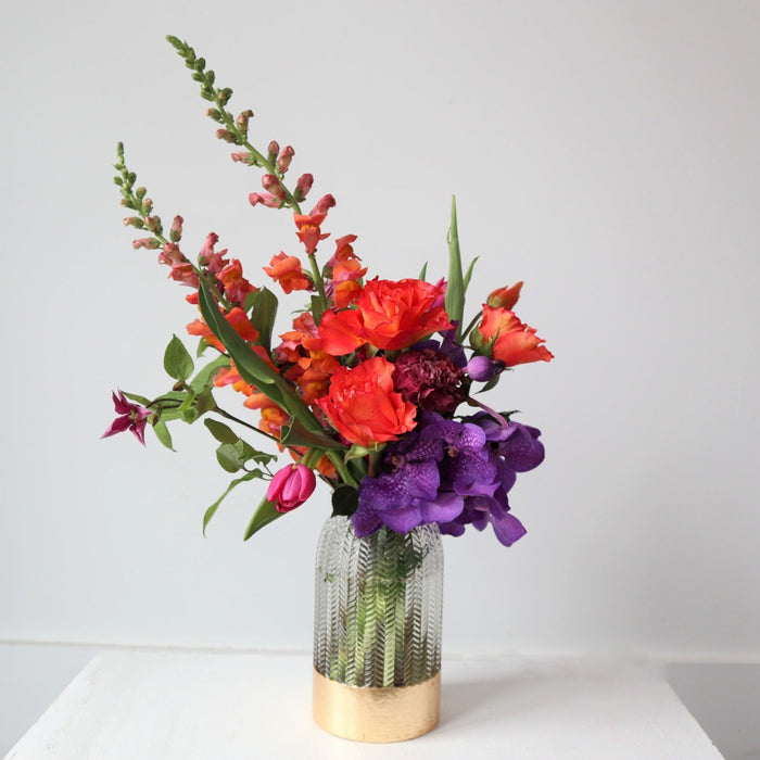White background with orange roses, orange snapdragons, purple vanda orchids, pink tulips designed in a clear vase with a gold rim bottom.