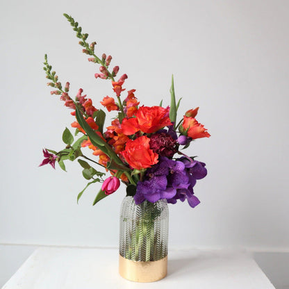 White background with orange roses, orange snapdragons, purple vanda orchids, pink tulips designed in a clear vase with a gold rim bottom.
